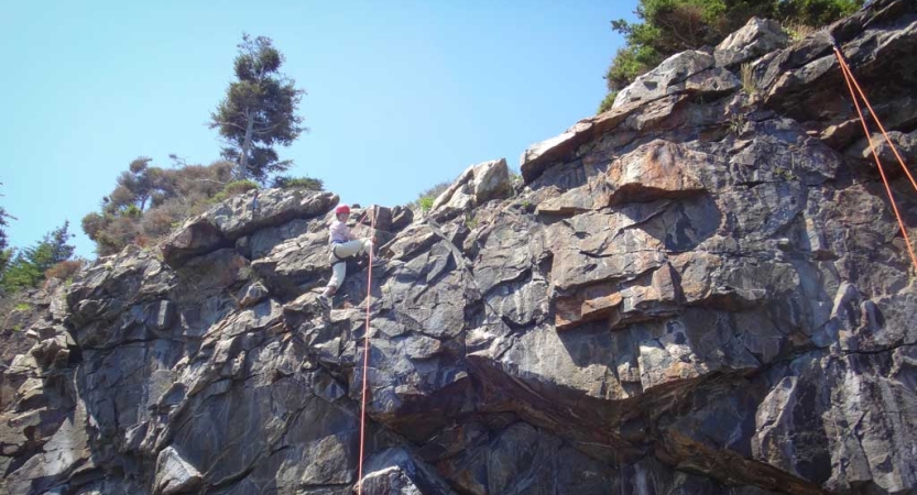 A person wearing safety gear is secured by ropes as they look down at the camera while rock climbing